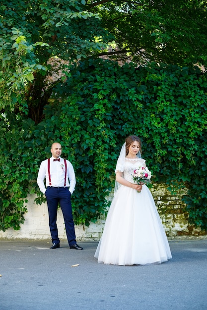 A noiva com um vestido branco em primeiro plano e o noivo com suspensórios atrás. na parede de fundo com folhas verdes