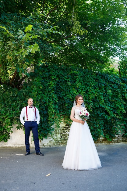 A noiva com um vestido branco em primeiro plano e o noivo com suspensórios atrás. na parede de fundo com folhas verdes