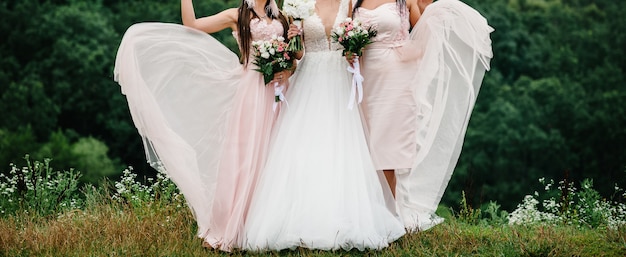 A noiva e as damas de honra em um vestido elegante estão de pé e segurando buquês de flores rosa pastel e verdes com fita na natureza. jovens garotas lindas segura um buquê de casamento ao ar livre.