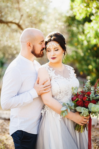 A noiva e o noivo com um buquê se abraçando entre as árvores em um bosque de oliveiras.