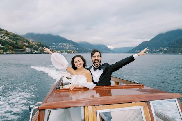 A noiva e o noivo sorrindo agitam as mãos enquanto estão de pé num iate a navegar no lago Como, na Itália.