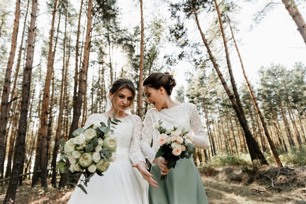 A noiva e sua amiga estão vestidas com vestidos de noiva e se divertem na floresta segurando buquês nas mãos. amizade feminina. dia do casamento. garotas se entregam