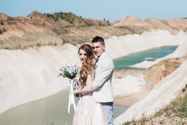 A noiva em um lindo vestido, abraçando o noivo em um terno claro perto do lago. Casal de noivos em pé em uma colina de areia ao ar livre. Uma história de amor romântica. Água azul-azulada no horizonte.