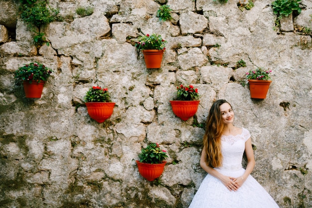 A noiva em um terno vestido de noiva fica perto da parede de um prédio antigo decorado com vasos de flores em perast