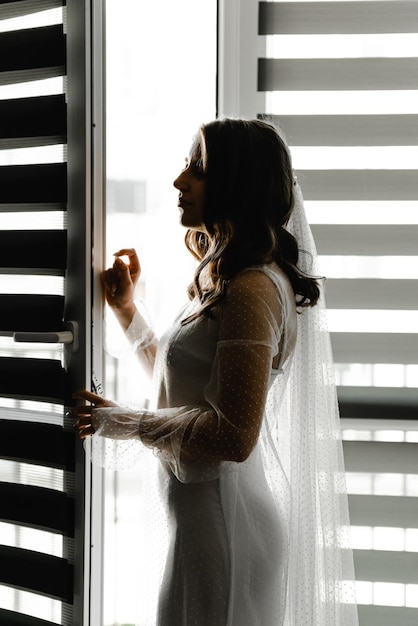 A noiva está esperando o noivo em uma sala com cortinas penduradas na janela. garota na janela. silhueta da noiva. persianas nas janelas. taxas do dia do casamento