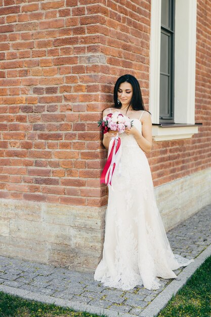 A noiva morena pensativa em lindo vestido de noiva mantém o buquê nas mãos, preocupa-se antes de uma ocasião especial em sua vida, fica perto do tijolo bilduing Modelo feminino com aparência perfeita