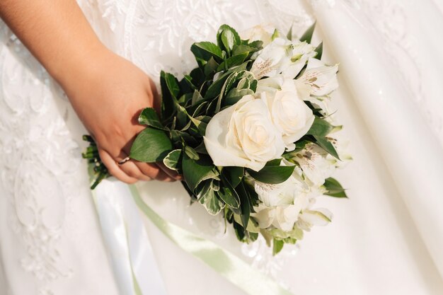 A noiva segura um buquê de rosas brancas no fundo de um vestido de noiva branco.