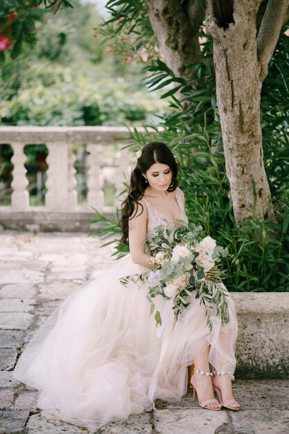 Foto a noiva sentada perto do oleandro em flor na cidade velha com um buquê de rosas brancas peônias e ramos de eucalipto em suas mãos