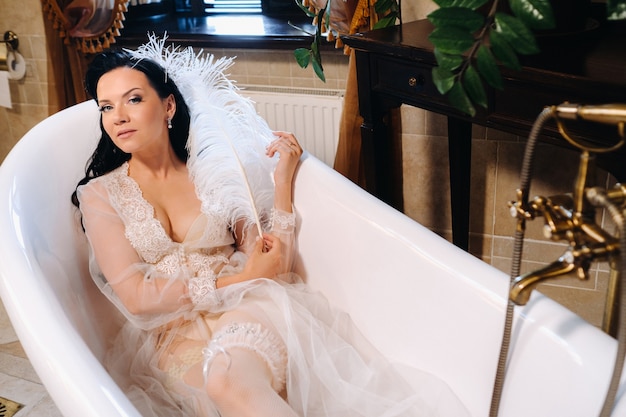 A noiva, vestida com um vestido boudoir transparente e calcinha, deita-se em um banheiro vintage com uma pena branca nas mãos.