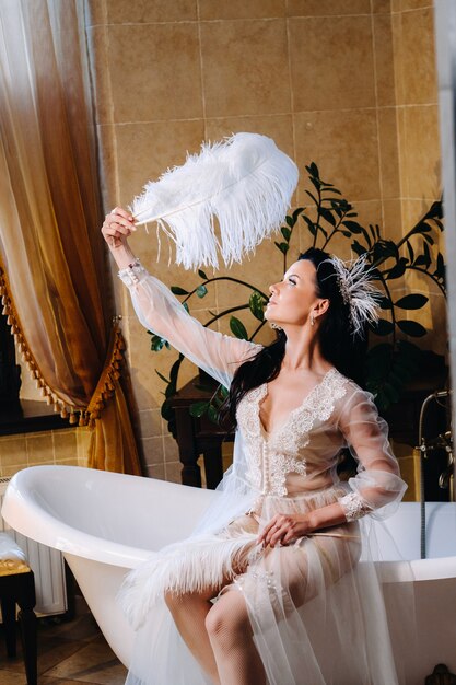 A noiva, vestida com um vestido boudoir transparente e calcinha, senta-se em uma banheira vintage com uma pena nas mãos, A manhã da noiva