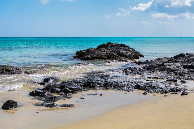 A paisagem tropical e paradisíaca de uma praia com areia loira e mar azul-turquesa com pedras negras.