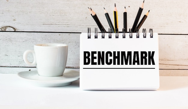 A palavra benchmark está escrita em um bloco de notas branco perto de uma xícara de café branca em uma parede de luz