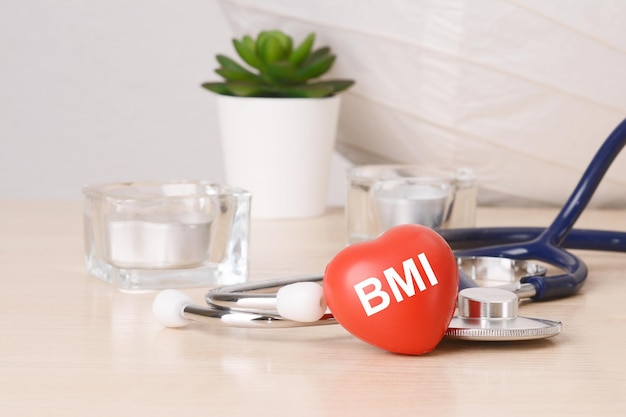 A palavra bmi está escrita em um brinquedo em forma de coração vermelho em uma mesa de madeira perto de um estetoscópio no fundo