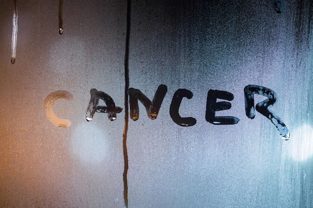 A palavra câncer escrita pelo dedo no vidro molhado noturno com luz de rua turva no fundo
