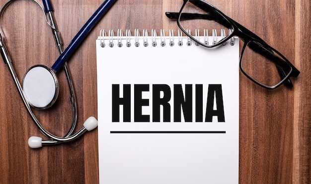 A palavra HÉRNIA está escrita em papel branco em uma parede de madeira perto de um estetoscópio e óculos de armação preta. Conceito médico