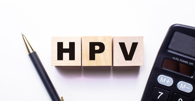 A palavra HPV é escrita em cubos de madeira entre uma caneta e uma calculadora em uma superfície clara