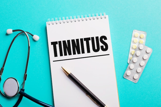 A palavra tinnitus escrita em um bloco de notas branco perto de um estetoscópio, canetas e comprimidos em uma superfície azul clara. conceito médico