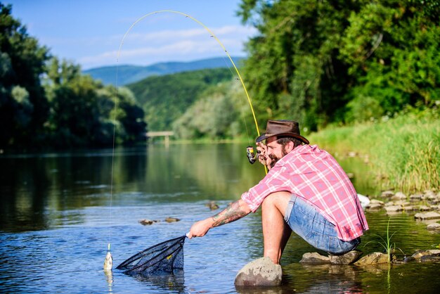 Foto a pesca do jogo grande relaxa na pesca da natureza hipster com hobbie de peixe voador de colher de homem hipster em camisa quadriculada pescador bem sucedido na água do lago homem barbudo maduro com peixe na vara