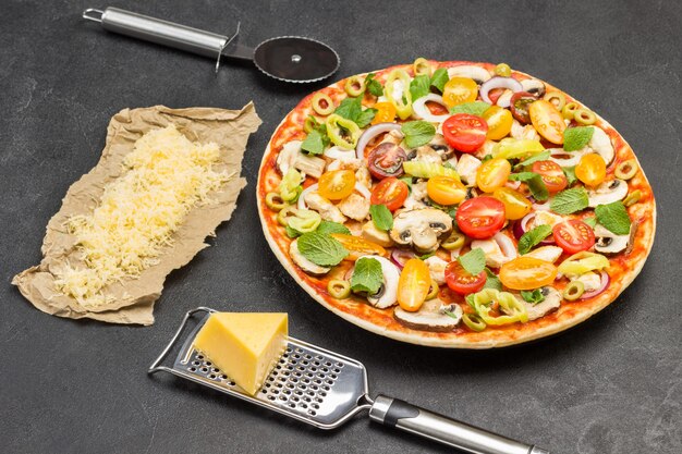 A pizza com ingredientes está pronta para assar. ralador de queijo na mesa. queijo ralado no papel. cortador de pizza. superfície preta. vista do topo