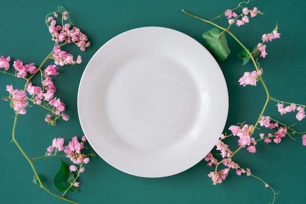 A placa vazia branca no fundo liso da tabela verde cercada pela pétala verde fresca floral e fresca cor-de-rosa lisa coloca, vista superior no prato de porcelana vazio limitado com flores cor-de-rosa e espaço da cópia gratuita.