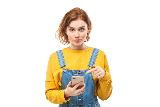 A ruiva alegre e chocada aponta o dedo para a tela em branco do smartphone em roupas amarelas isoladas no fundo branco do estúdio