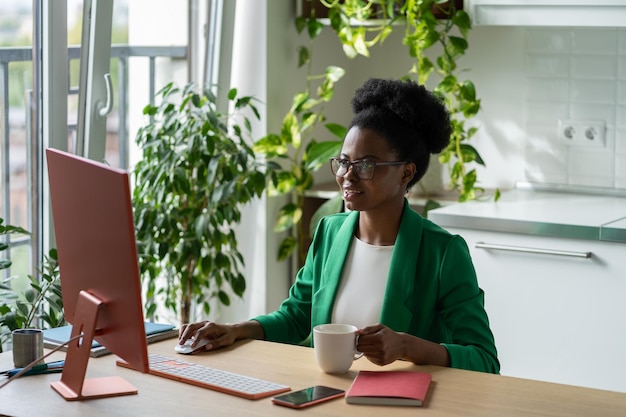 A secretária da jovem afro-americana trabalhando senta-se na mesa do escritório com o computador e bebendo café