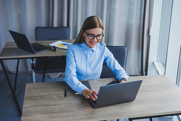 A secretária verifica a agenda uma jovem alegre e bonita de óculos com um sorriso usa um laptop enquanto está sentada em seu local de trabalho