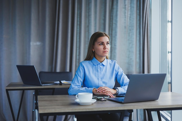 A secretária verifica a agenda uma jovem alegre e bonita de óculos com um sorriso usa um laptop enquanto está sentada em seu local de trabalho