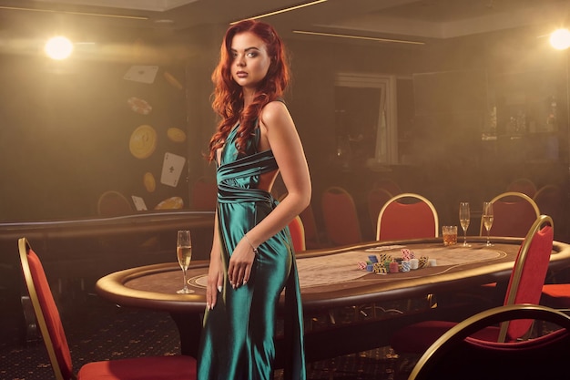 A sedutora senhora ruiva em um longo vestido de cetim azul está posando de lado contra uma mesa de pôquer no cassino de luxo. Paixão, cartas, fichas, álcool, vitórias, jogos de azar - é como entretenimento feminino. Fundo de fumaça