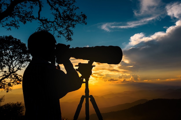 A silhueta de um fotógrafo da paisagem usa a lente teleobjetiva super no alto das montanhas durante o por do sol com o céu do por do sol de dois tons