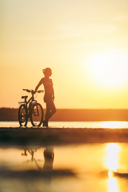 A silhueta de uma garota esportiva em um terno de pé perto de uma bicicleta na água ao pôr do sol em um dia quente de verão. conceito de aptidão.