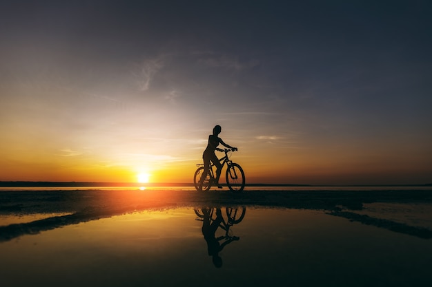 A silhueta de uma garota esportiva em um terno que se senta em uma bicicleta na água ao pôr do sol em um dia quente de verão. Conceito de aptidão.