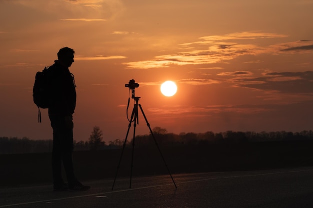 A silhueta de uma pessoa contra o fundo do sol e o lindo céu O contorno de um homem ao lado de um tripé e uma câmera no fundo do pôr do sol