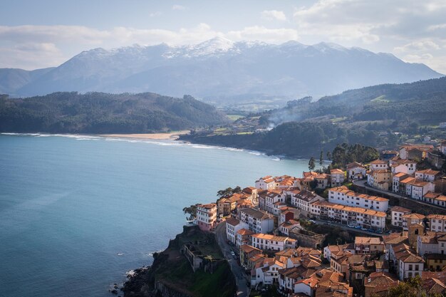 A tomada horizontal captura o charme costeiro de Lastres, picos nevados e águas azuis.