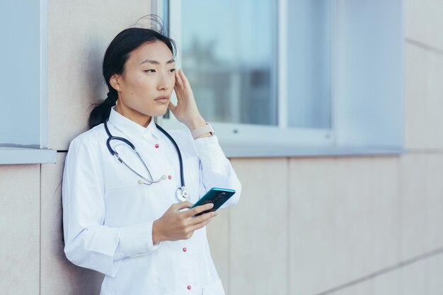 A triste enfermeira asiática, perto da clínica durante uma pausa, usa o telefone, a mulher tem uma forte dor de cabeça, excesso de trabalho deprimido