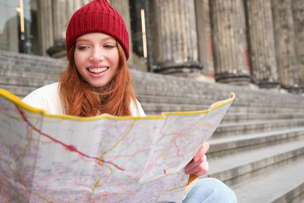 A turista sorridente se senta na escada perto do museu e segura o mapa de papel explora a cidade durante o si