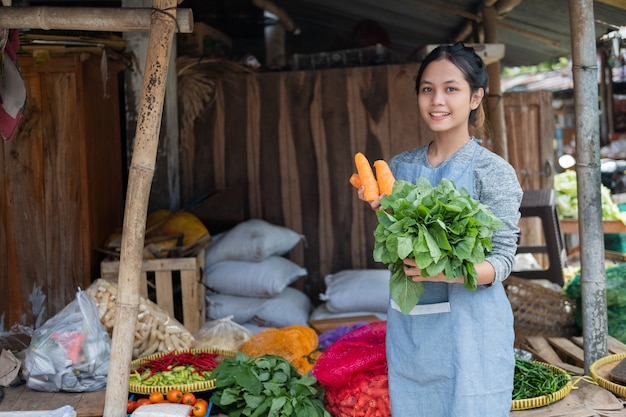A verdureira asiática sorri enquanto segura cenoura e espinafre em uma barraca de legumes em um mercado tradicional