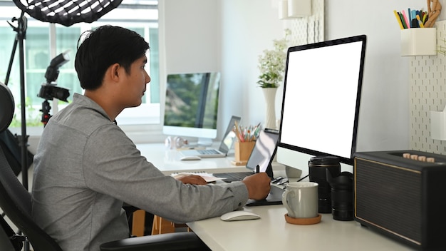 A visão lateral do designer gráfico ou fotógrafo está usando a mesa gráfica para retocar uma foto em seu espaço de trabalho.