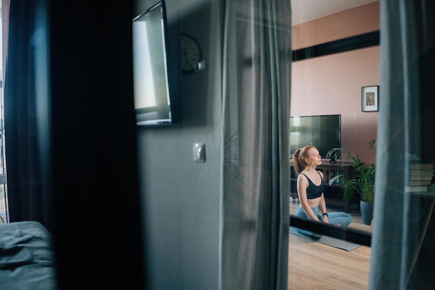 A visão remota da jovem ruiva vestindo roupas esportivas está meditando em posição de lótus sentada no tapete de ioga perto do laptop. Senhora ruiva pacífica segurando as mãos no joelho na posição Om.