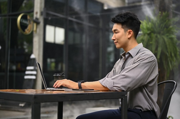 A vista lateral de um homem está usando seu laptop e sentado em uma cafeteria em um espaço ao ar livre