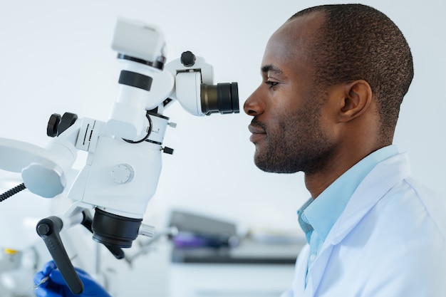 A vista lateral de um jovem dentista bonito usando um microscópio profissional e confirmando seu diagnóstico