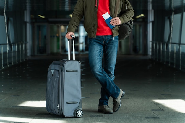 A vista recortada do turista masculino está pronta para a viagem. Ele está de pé no aeroporto e carregando sua mala. Homem está segurando seu passaporte