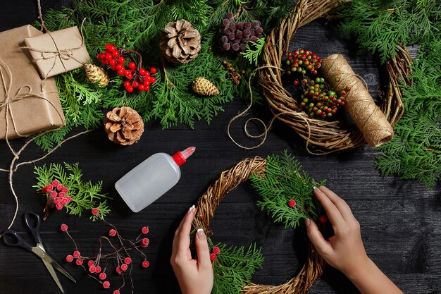 A vista superior de mãos femininas faz uma guirlanda de Natal embalada em presentes e pergaminhos de ramos de abeto e ferramentas ...