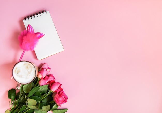 A xícara de café quente no bloco de notas vazio do café da manhã com caneta engraçada e buquê de rosas frescas em fundo rosa suave