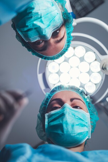 Desde abajo, cirujanas con uniforme médico usando herramientas profesionales mientras están de pie bajo un brillante l