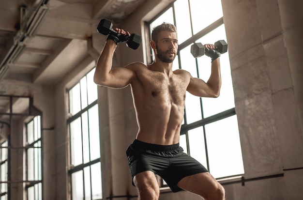 Desde abajo, fuerte atleta masculino en cuclillas con pesas pesadas durante el entrenamiento funcional en el gimnasio