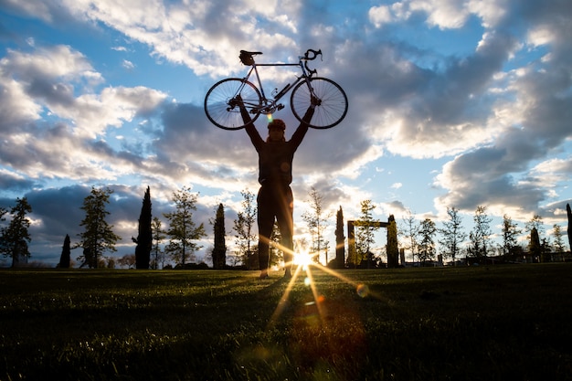 Desde abajo joven feliz guapo macho levantando las manos con bicicleta en el parque de la ciudad en la puesta de sol en la parte posterior iluminada