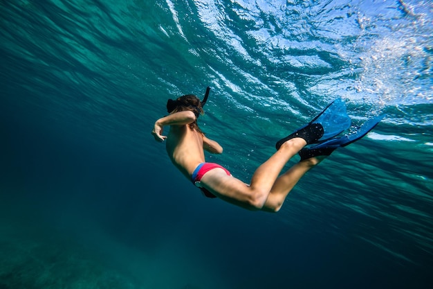 Desde abajo de todo el cuerpo adolescente anónimo con máscara de buceo y aletas buceando bajo el agua en el mar azul profundo