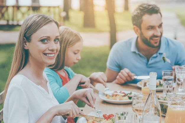 Abendessen mit den Nächsten genießen. Glückliche Familie, die zusammen Essen genießt, während die Frau in die Kamera schaut und lächelt