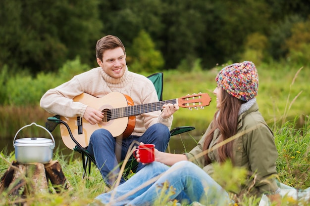 Abenteuer-, Reise-, Tourismus- und Personenkonzept - lächelndes Paar mit Gitarre, das Essen kocht und beim Camping trinkt